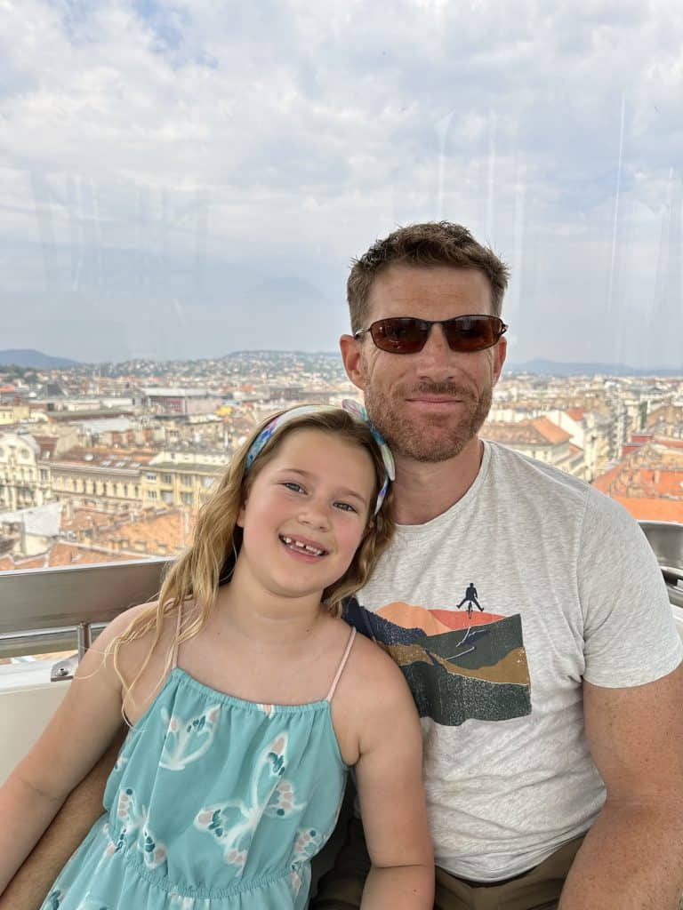 Mr Tin Box and our eight-year-old daughter smiling at the camera while sat in a capsule on the Budapest Eye. There's a view of the city in the background