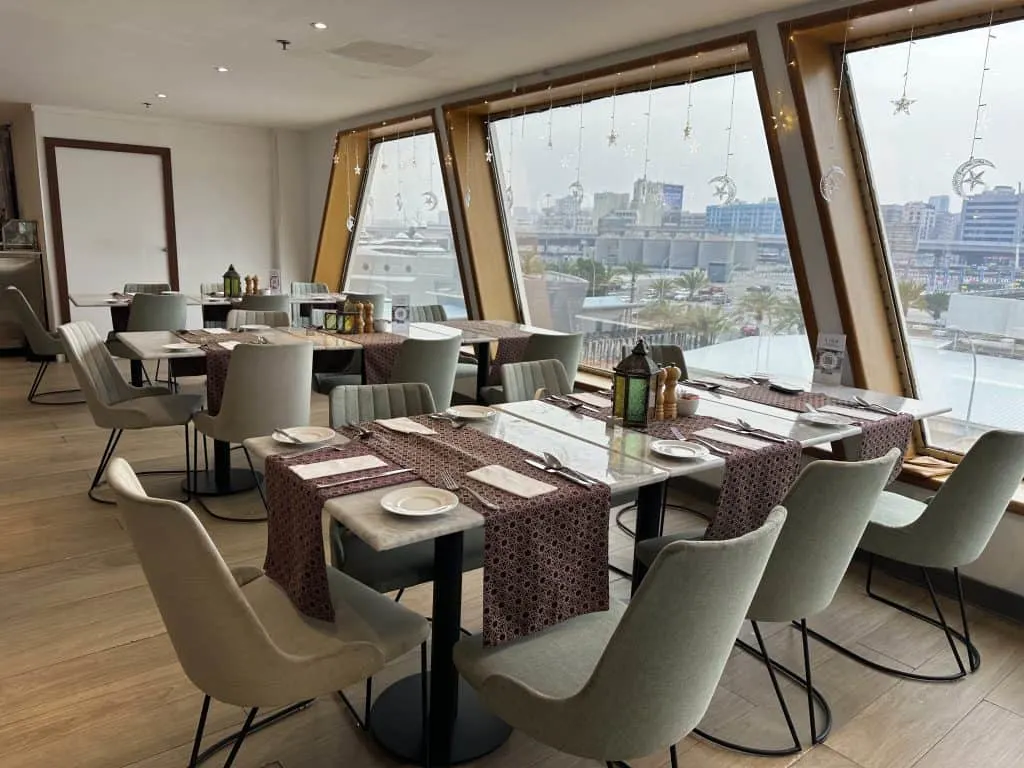 Restaurant tables laid out in the Lido onboard QE2. There is a view of Dubai out of the floor to ceiling windows