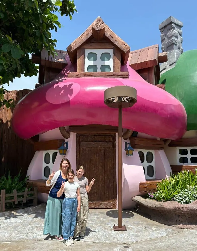 Claire and her daughters doing a peace sign in front of a giant Smurf house in the Motiongate theme park