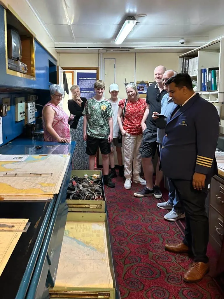 QE2 ship's tour guide showing guests the chartroom behind the ship's bridge. Everyone is smiling at an open draw of old electrical equipment that was the bridge's junk draw