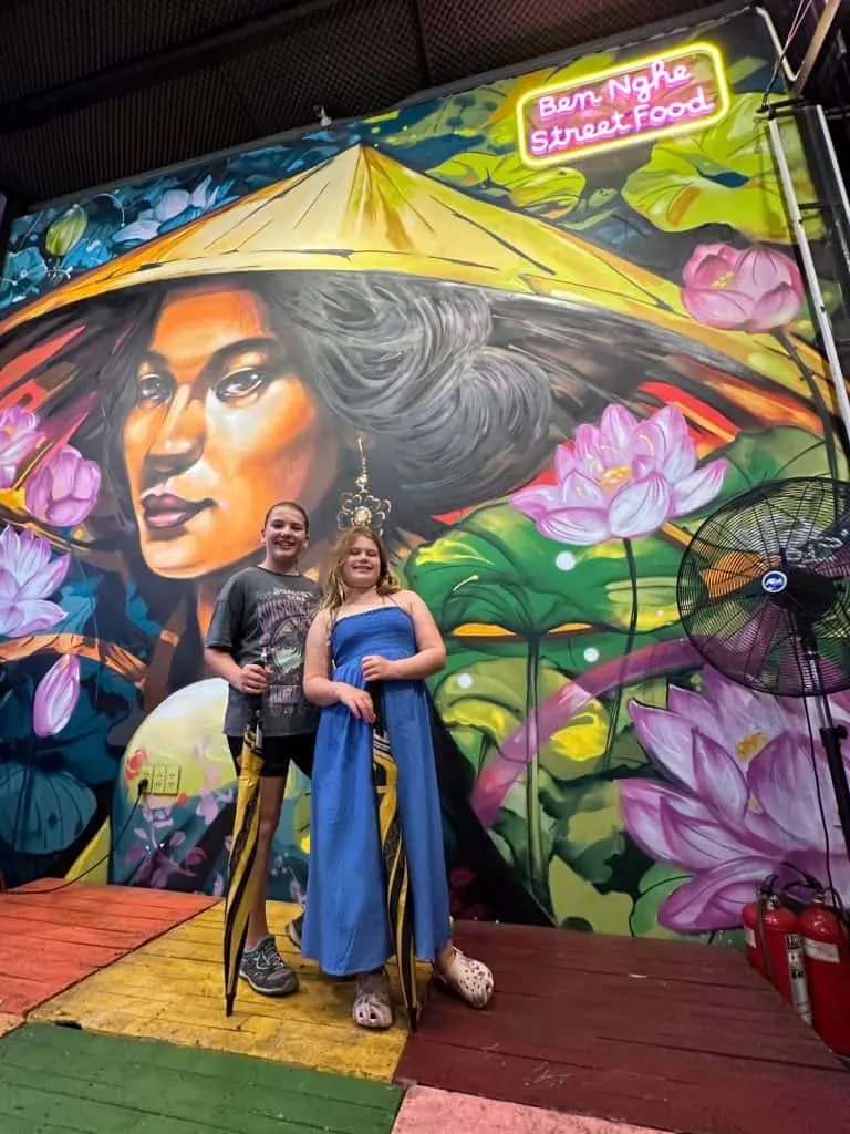 Our daughters stood in front of a colourful mural in the Bến Nghé Street Food Market