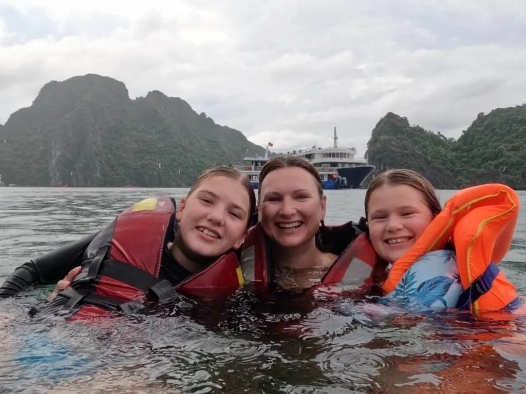 Claire and her daughters in the water at Ha Long Bay. They are wearing life jackets and there is a cruise ship behind them