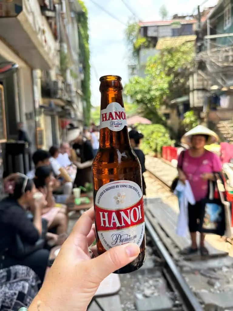 My hand holding a bottle of Hanoi beer with a view of Train Street behind