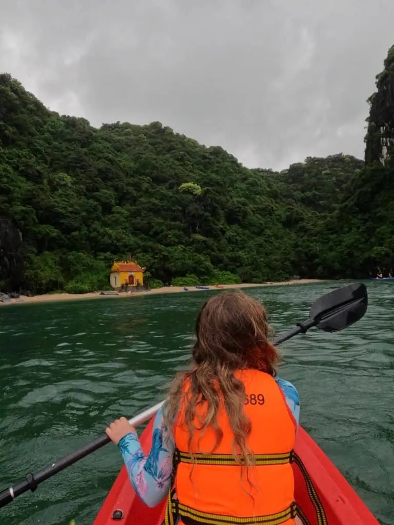 Our nine-year-old daughter paddling a kayak with the mountains of Ha Long Bay and a yellow fisherman's temple in front of her