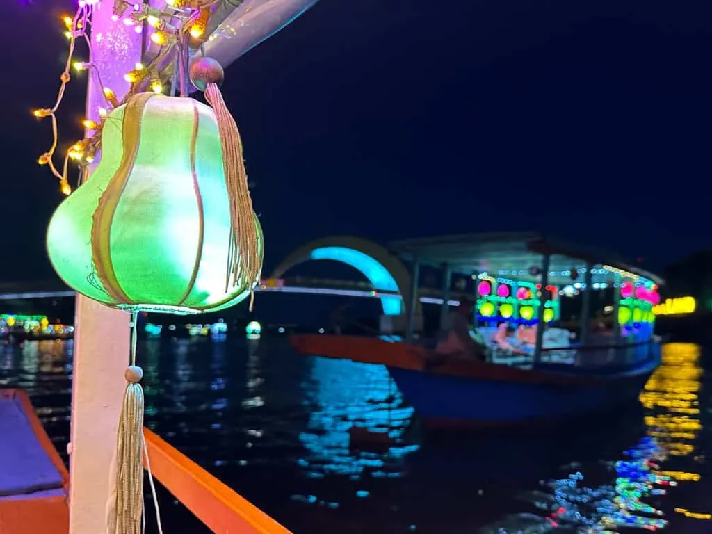 A green lantern hanging off the side of a boat at night. In the background there's a lantern boat and the Moon Bridge in Hoi An