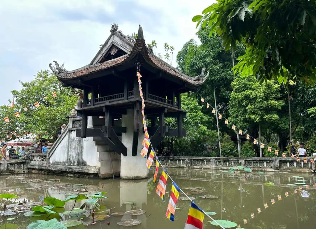 The One Pillar Pagoda which is stood in a pond with Buddhist flags handing from the rafters to the edge of the pond