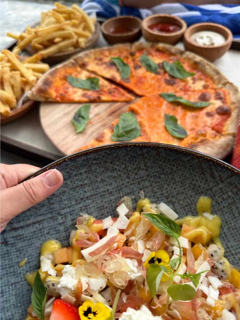 A plate of finely sliced fruit salad and a pizza in the background