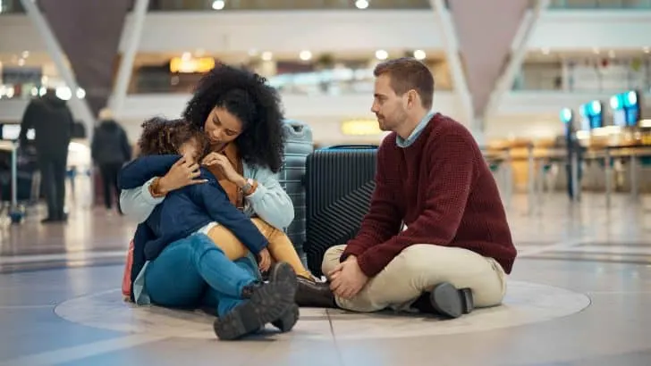 Family sat on an airport floor. The child is sleeping in her mother's arms. They all look sad like something has gone wrong