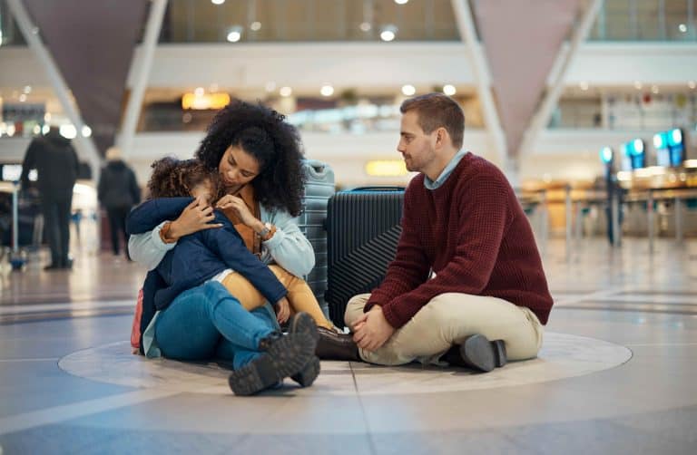 Family sat on an airport floor. The child is sleeping in her mother's arms. They all look sad like something has gone wrong