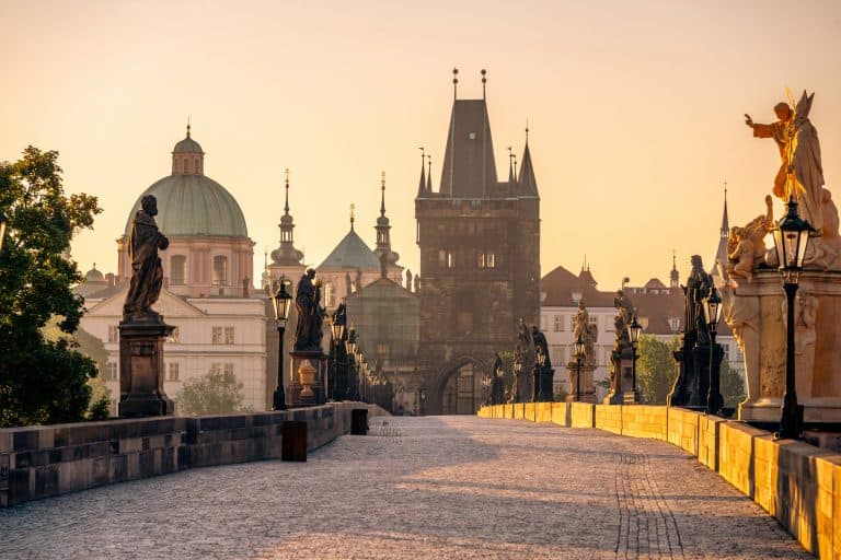Sunrise casts a warm glow over Charles Bridge in Prague, highlighting the statues and the historic architecture. The Vltava River glistens, creating a serene morning atmosphere.