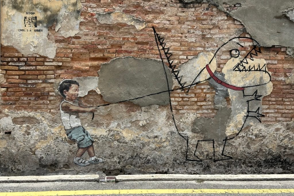Street art in Penang showing a young boy holding back a cartoon dinosaur on a leash