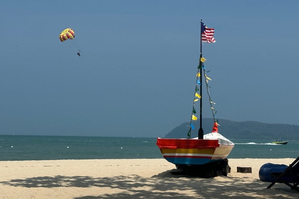 A paraglider is pulled along by a speed boat in the sea in front of Holiday Villas Langkawi. A small, colourful sailing boat is on the beach in the foreground. It is flying the Malaysian flag