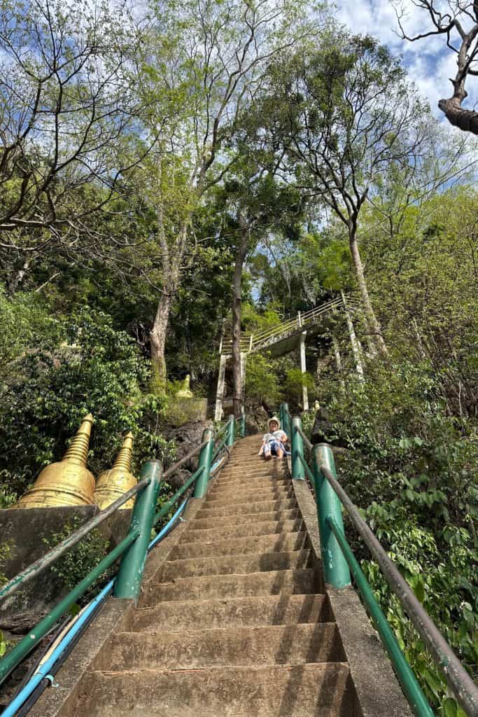 A column of steps going up towards the Tiger Cave in Krabi. There are golden bells on the left and my daughter is sat on the steps part way up. They seem to go on forever