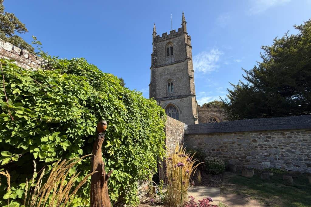 A church seen over the wall of Avebury Manor gardens with a small owl sculpture by Martin Adamson in the foreground