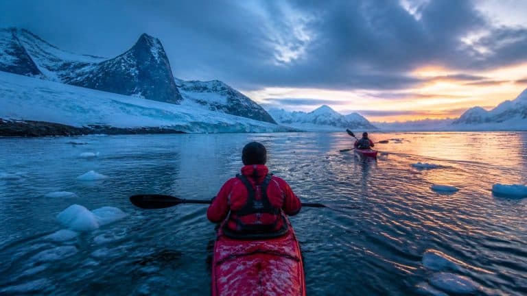 Adults wearing warm winter clothing and drysuits kayaking through icy waters off South Georgia, with a typical South Georgia coastal landscape in the background - rugged dark mountains, glaciated slopes