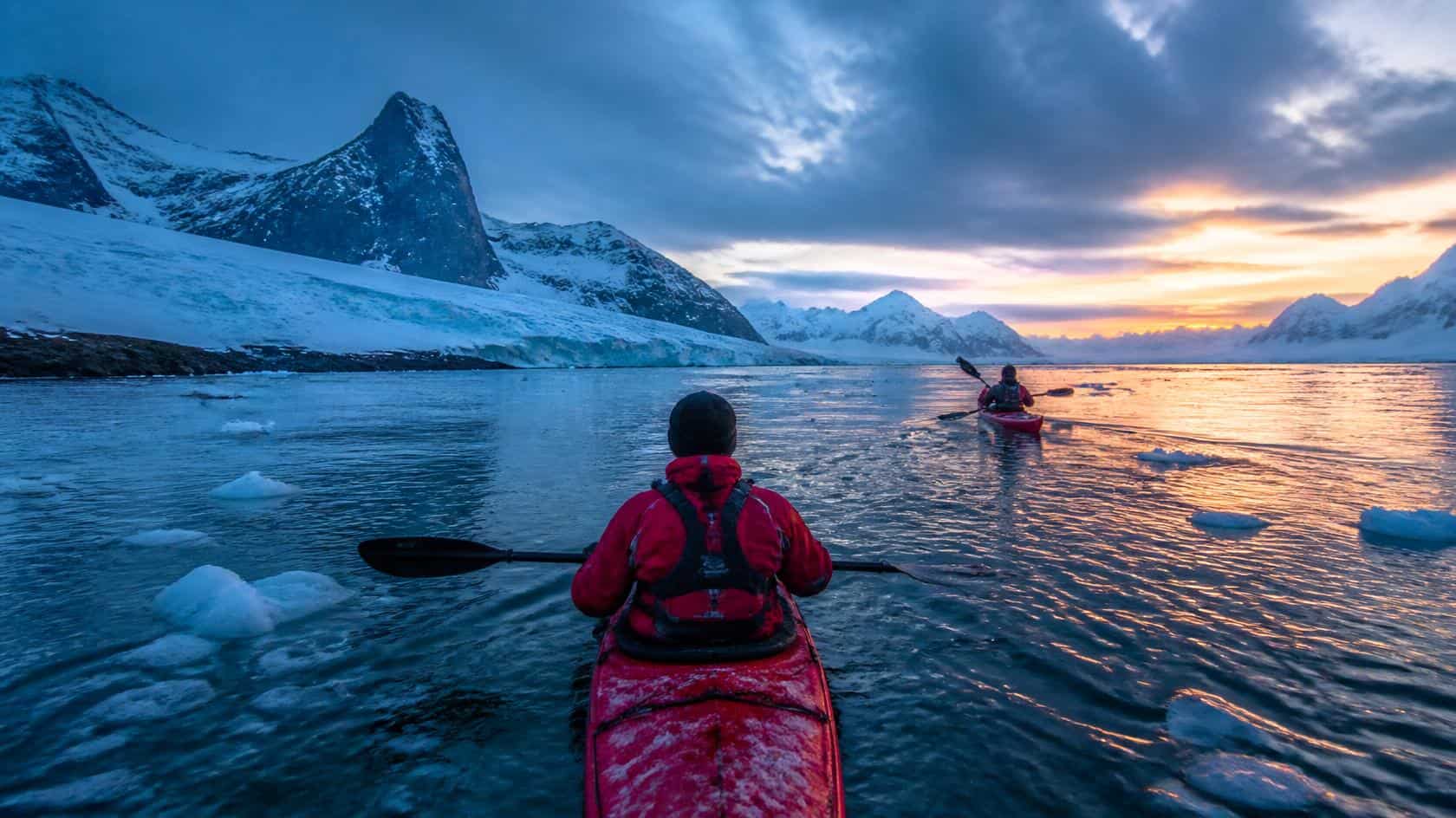 Adults wearing warm winter clothing and drysuits kayaking through icy waters off South Georgia, with a typical South Georgia coastal landscape in the background - rugged dark mountains, glaciated slopes